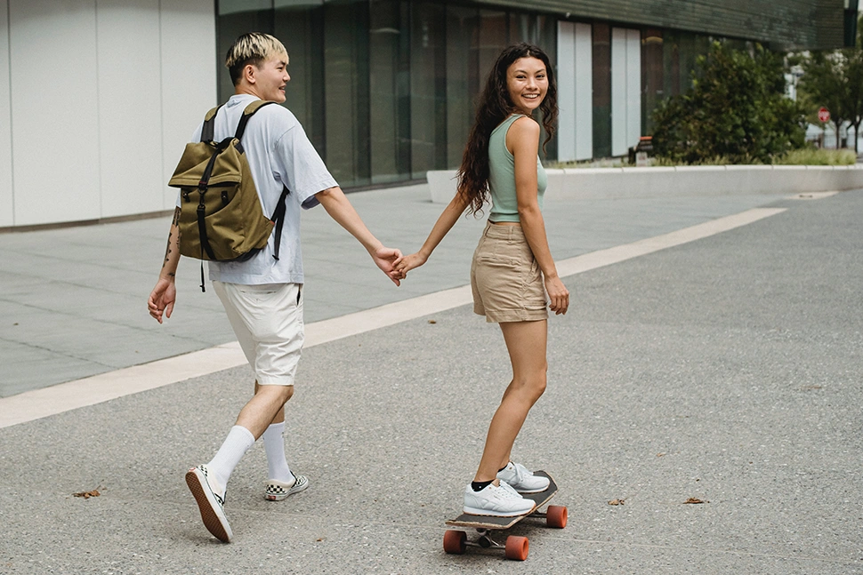 Woman on a skateboard holding hands with a man as he walks next to her, she is looking back smiling at the camera while he is smiling at her