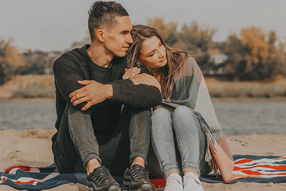 Man and woman sitting on a beach, she is using a blanket to cover her and leaning into his shoulder for support