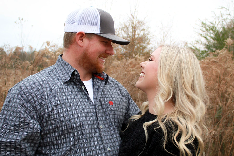 Man and woman in a field together smiling at one another, he is wearing a hat