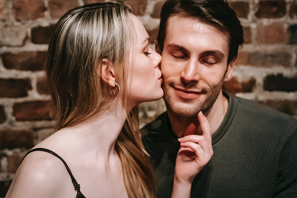 Woman kissing a mans cheek as he closes his eyes and smiles, she is bringing him closer with her hand