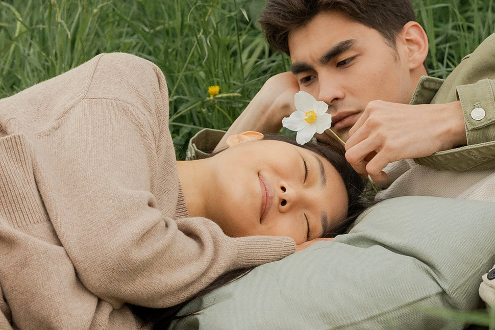 Man and woman are laying together in a grass field, he is holding a flower looking at her and she is smiling leaning into his chest