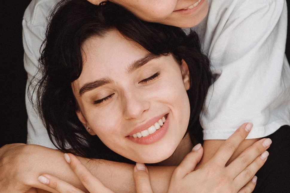 Woman being hugged from behind, she has her eyes closed and is smiling